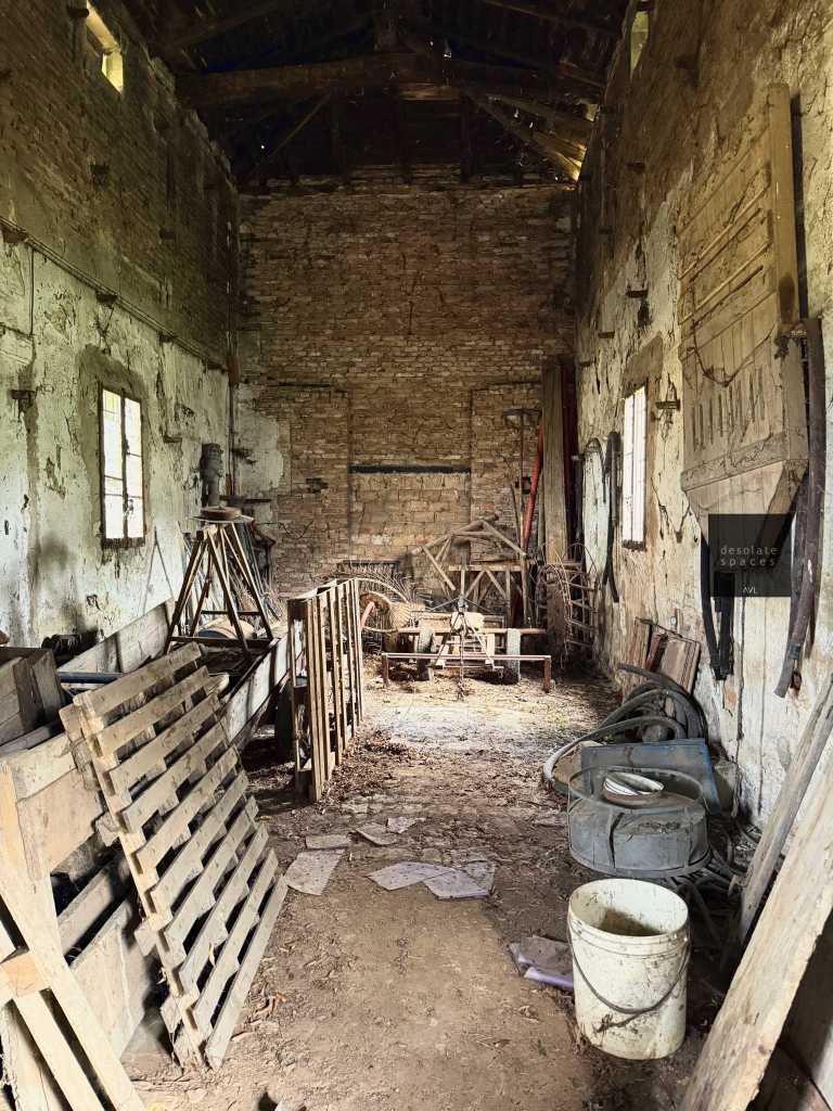 Interno di un vecchio magazzino con muri di mattoni e attrezzature agricole abbandonate, come pallet di legno e un secchio bianco.