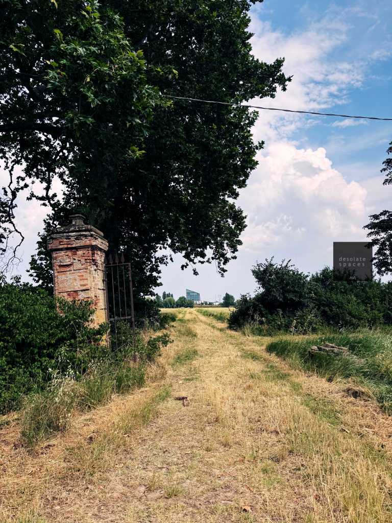 Sentiero rurale che conduce tra alberi e cespugli, con un cancello in mattoni visibile a sinistra e cielo nuvoloso sullo sfondo.