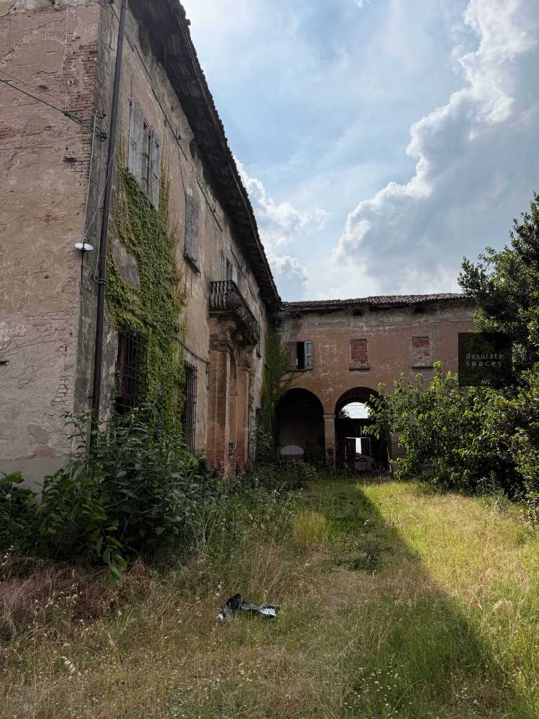 Facciata di un edificio abbandonato con pareti scrostate e vegetazione che cresce attorno.