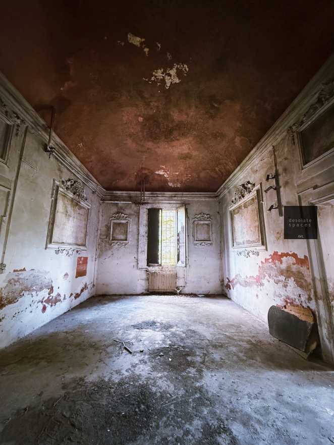 Interno di una stanza abbandonata con pareti danneggiate e soffitto scrostato. Una finestra sul fondo offre un'illuminazione tenue.