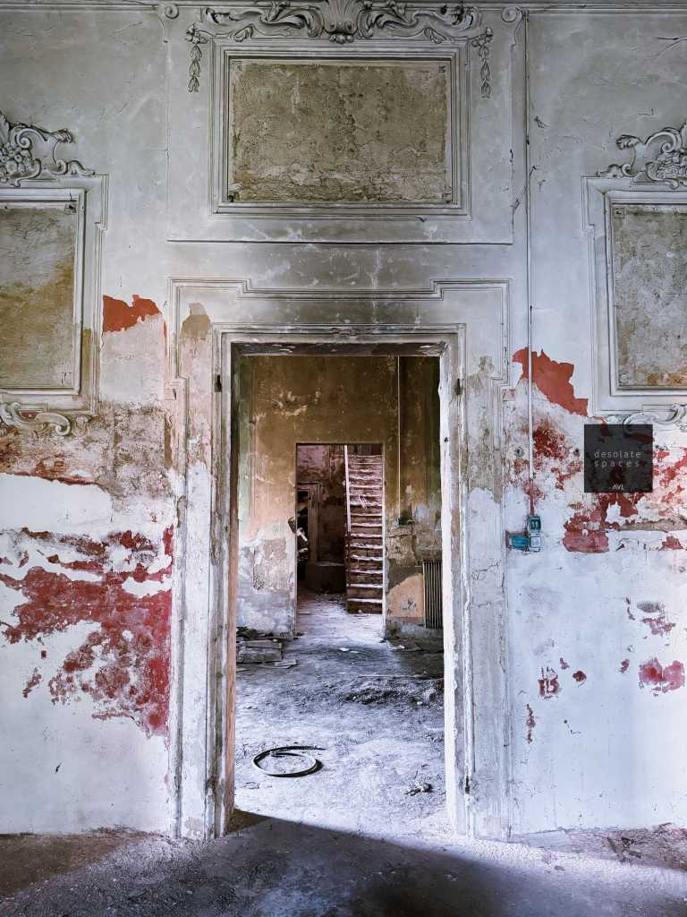 Una porta aperta che conduce a un interno trascurato con pareti scrostate e segni di umidità, una scala di legno visibile sullo sfondo.