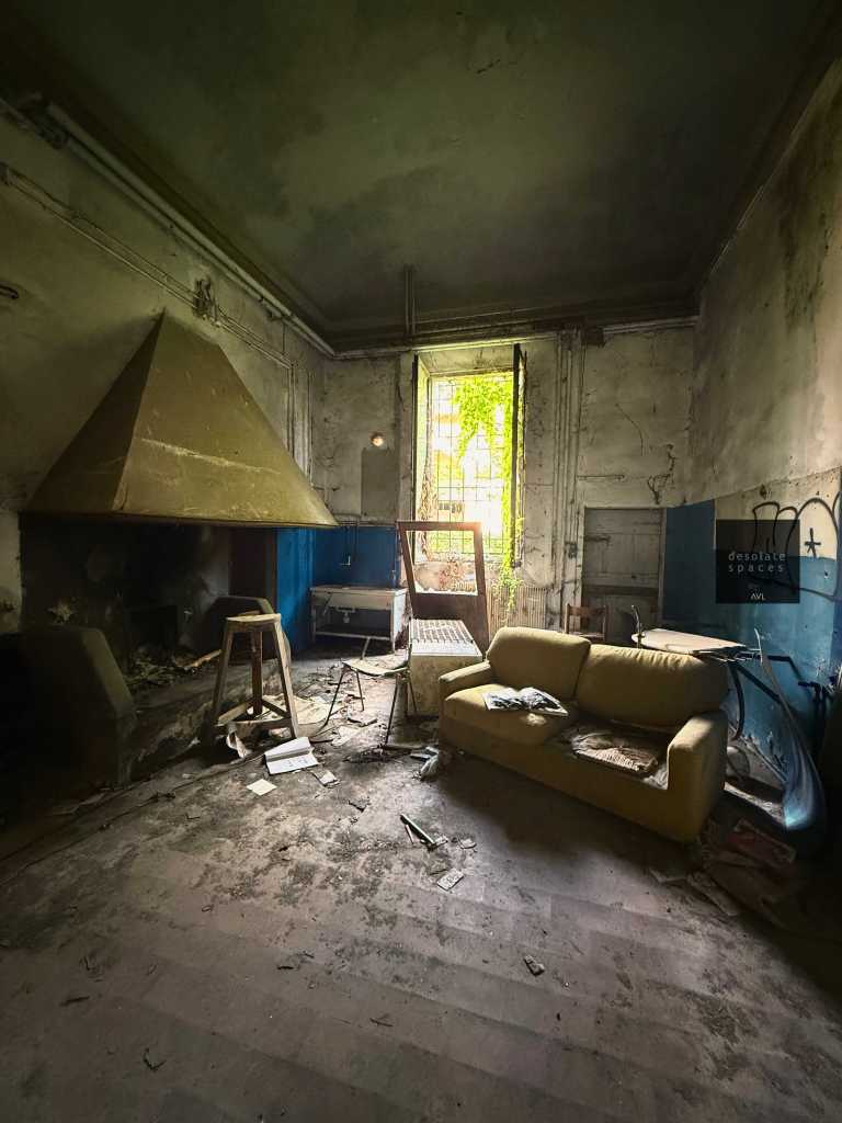 Interno di una stanza abbandonata con un camino, un divano sporco e un'atmosfera di degrado. La luce entra attraverso una finestra aperta su di un giardino incolto.