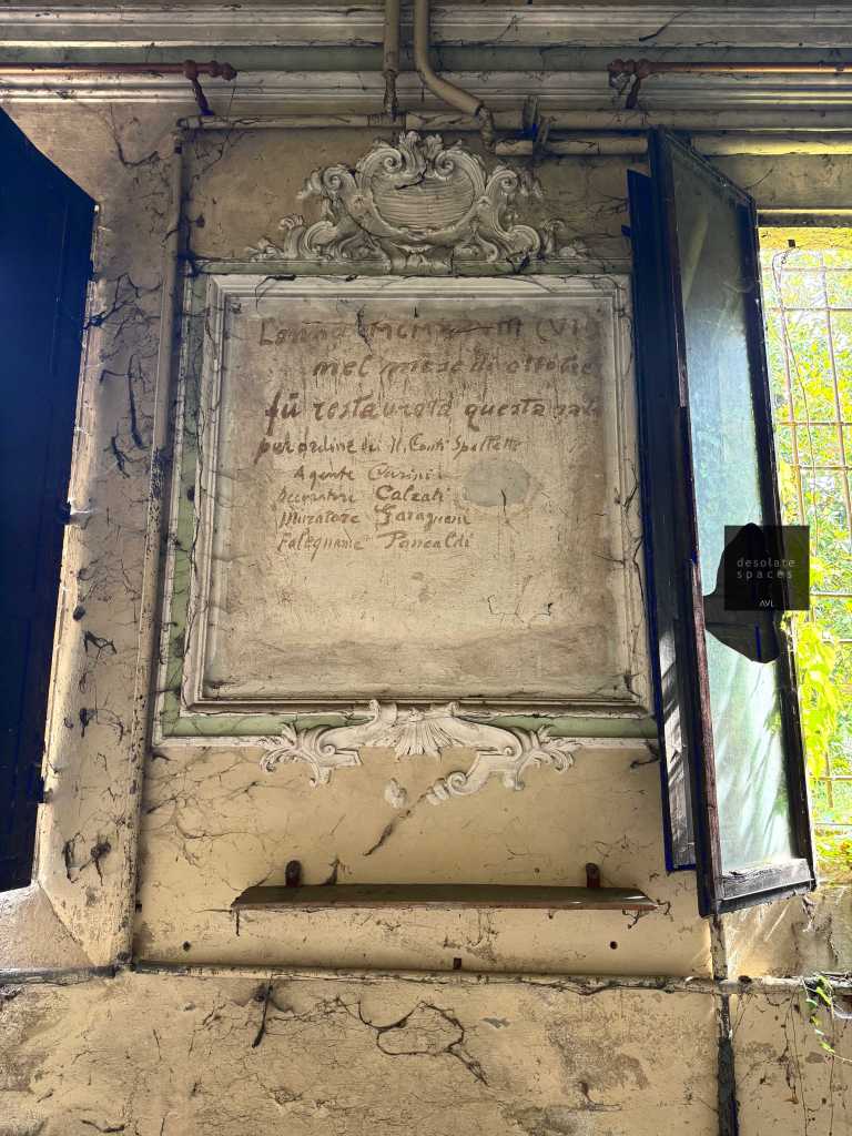 Una parete con una cornice decorativa contenente un testo scritto, esposta vicino a una finestra aperta che lascia entrare la luce, in un ambiente con muri crepati.