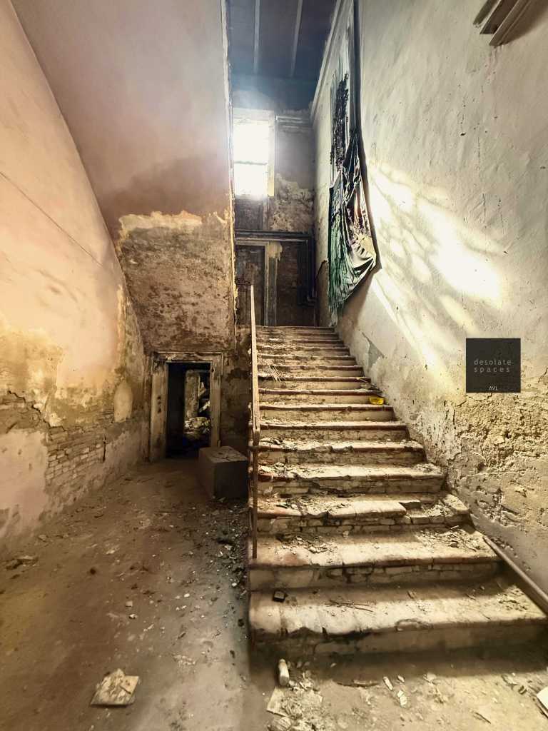 Scalinata sporca e abbandonata in un ambiente deteriorato, con pareti scrostate e luce che entra da una finestra in cima.