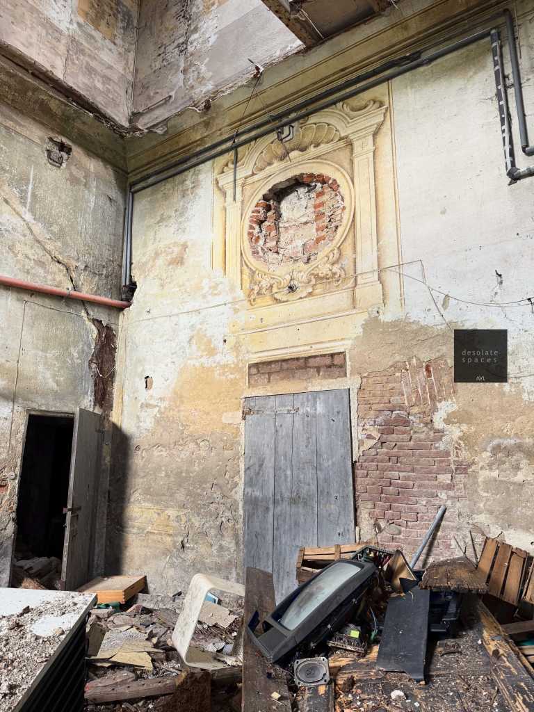 Interno di un edificio abbandonato con pareti danneggiate e un affresco parzialmente visibile. Ci sono detriti e oggetti sparsi sul pavimento, inclusi mobili rotti e un vecchio televisore.
