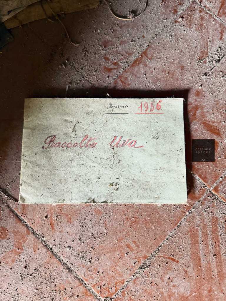 Un libro usato con la scritta 'Raccolto Uva' e l'anno '1956' su una copertina di carta, posato su un pavimento polveroso.