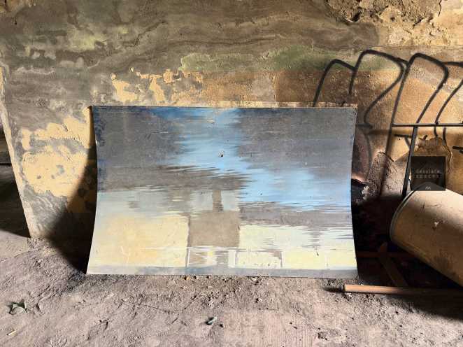 Una tavola dipinta con un riflesso blu e giallo, appoggiata su un pavimento di terra con pareti grezze sullo sfondo.