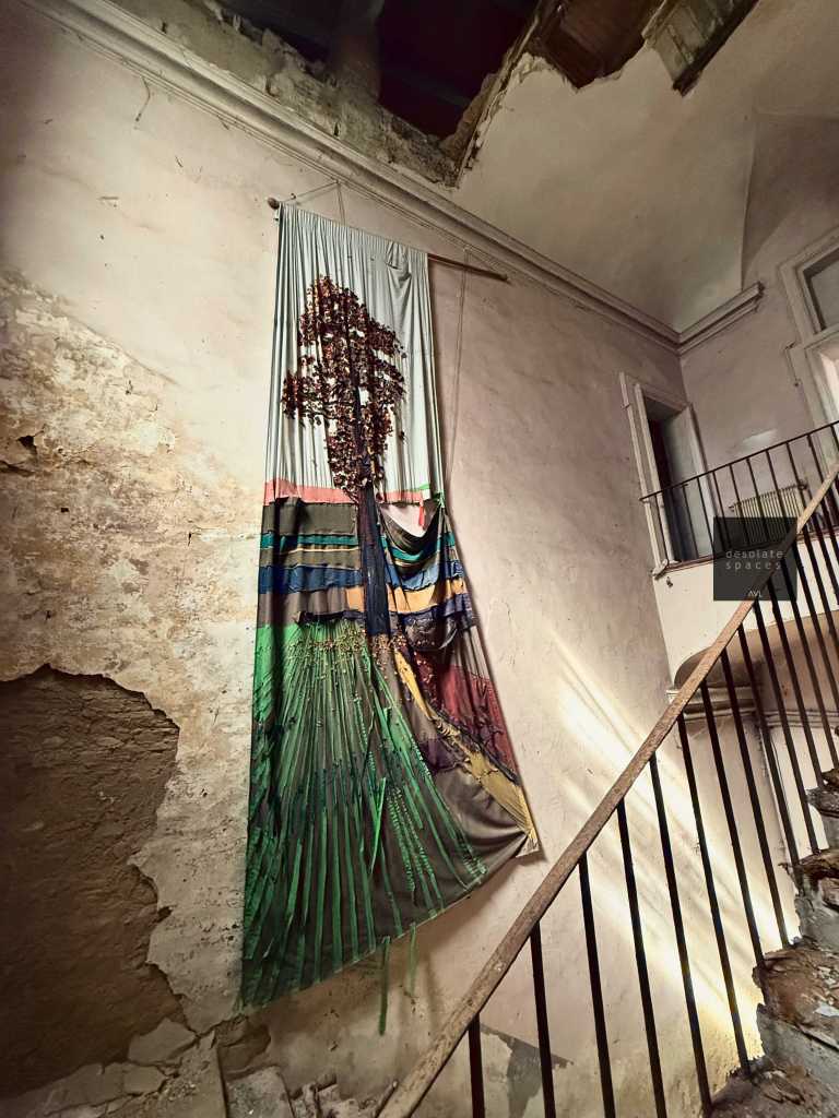 Arazzo appeso su un muro di una scala in un edificio abbandonato, con un design che rappresenta un albero e paesaggi colorati.
