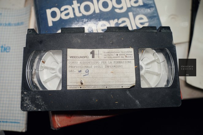 Un nastro VHS con etichetta, intitolato 'Corso audiovisivo per la formazione professionale degli infermieri', con evidenti segni di usura, posizionato su una scrivania accanto a un libro di patologia generale.