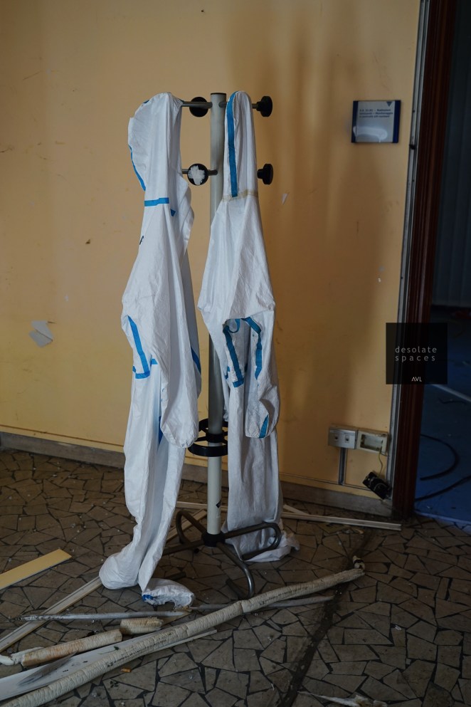 Due tute protettive bianche appese a un appendiabiti in una stanza vuota con pareti gialle.
