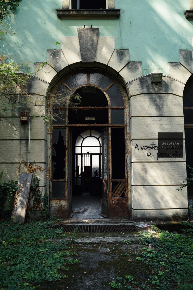 Entrata di un edificio abbandonato con pareti verdi e una porta arrugginita. Scritta graffiti sulla parete: 'A vostro piacere e per amor'. Vegetazione che cresce attorno all'ingresso.