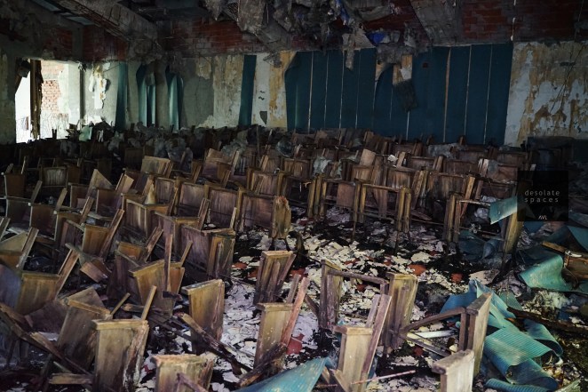 Interno di una sala abbandonata con molte sedie disposte in modo disordinato, circondate da detriti e segni di degrado.