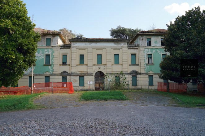 Facciata di un edificio abbandonato con pareti verdi e finestre chiuse, circondato da alberi e recinzione arancione.